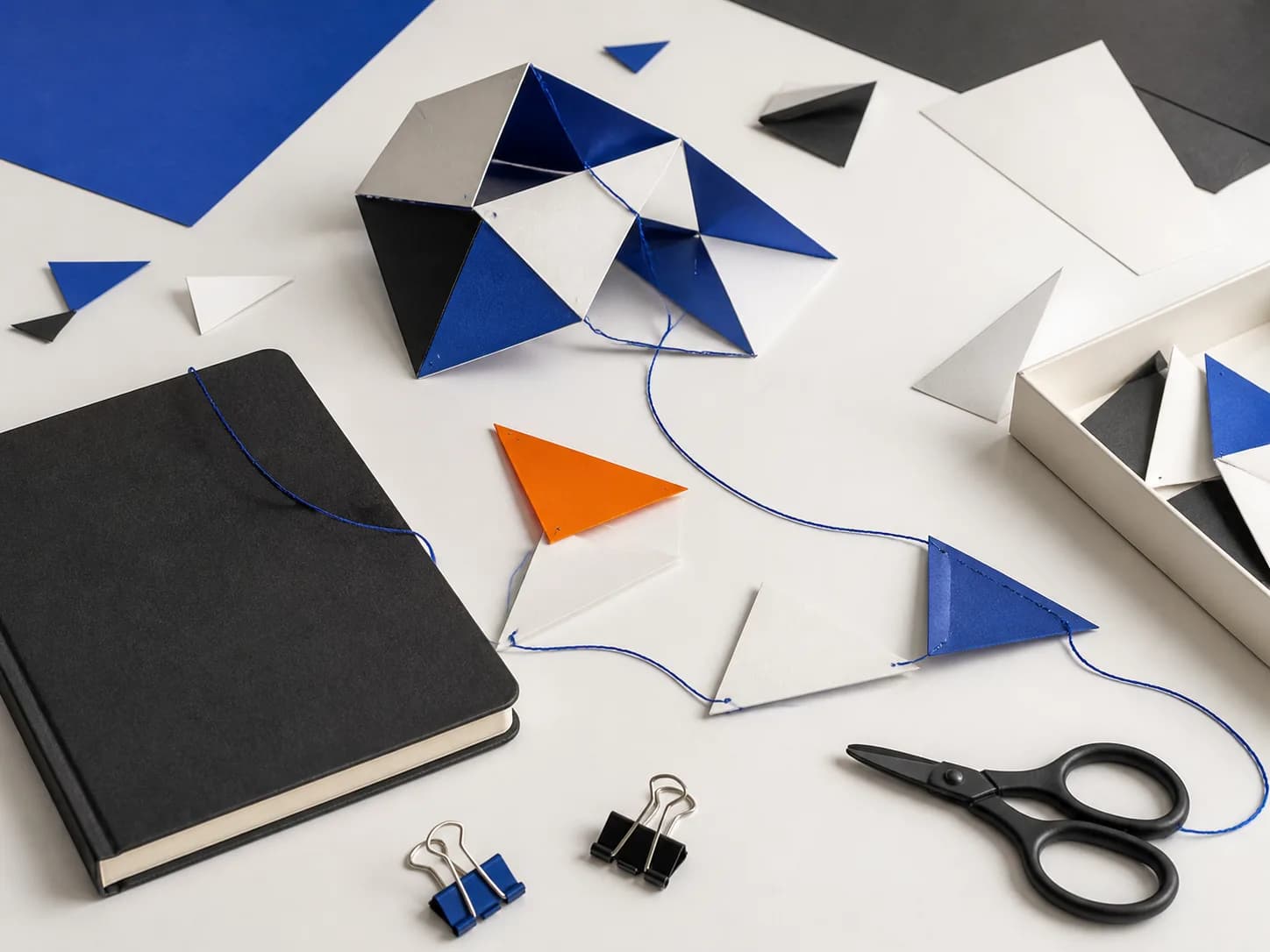 An unfinished paper construction links a notebook, folded pieces, thread, clips, and scissors on a bright table.